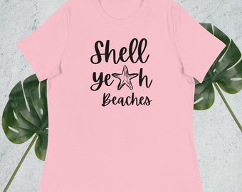 May include: A pale pink t-shirt featuring the phrase "Shell yeah Beaches" in black script, with a starfish graphic in the word "yeah". The shirt has a relaxed fit, suitable for beach outings or everyday wear.