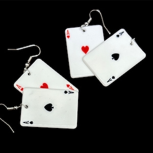 May include: A pair of earrings featuring white playing card charms with black and red suit symbols. The charms are shaped like playing cards with the letter 'A' and a heart or spade symbol.
