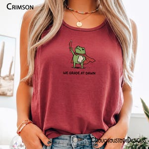 May include: A crimson tank top featuring a graphic of a green frog wearing a red cape, holding a red pen and a paper with an A+ grade. The text below the frog reads WE GRADE AT DAWN. The shirt has a round neckline and a sleeveless design.