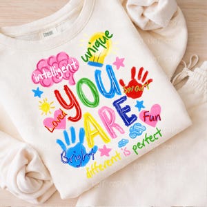 May include: A cream-coloured sweatshirt featuring the words "YOU ARE" in vibrant, handwritten lettering. The design incorporates the words "intelligent," "unique," "loved," "smart," "fun," "bright," and "different is perfect," alongside various illustrations.