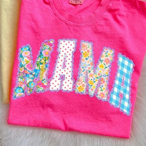 May include: A vibrant pink t-shirt featuring the word "MAMA" in appliqued letters. The letters are adorned with floral prints, polka dots, and a blue gingham design, all edged with a scalloped trim.