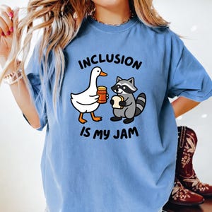 May include: A light blue t-shirt with the text "INCLUSION IS MY JAM". The graphic features a white goose holding a jar of jam and a gray raccoon holding a piece of bread. The shirt is a comfortable, casual style.