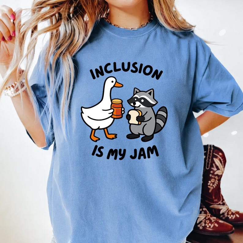 May include: A light blue t-shirt with the text "INCLUSION IS MY JAM". The graphic features a white goose holding a jar of jam and a gray raccoon holding a piece of bread. The shirt is a comfortable, casual style.