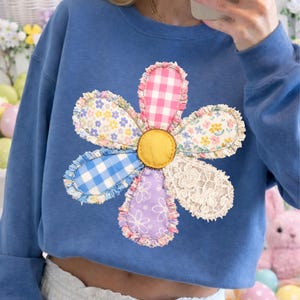 May include: Blue sweatshirt featuring a large, colorful flower applique. The flower has a yellow center and petals in various patterns, including gingham, floral, and lace. The sweatshirt is a solid blue color.