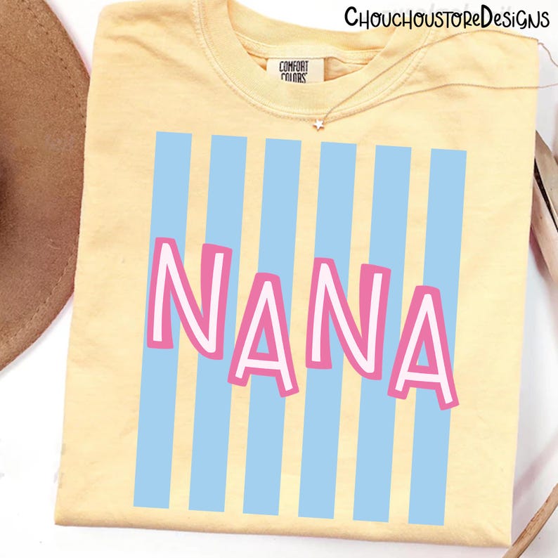 Puede incluir: Una camiseta amarillo claro con la palabra "NANA" en letras rosas con contorno blanco, sobre un fondo de rayas verticales azul claro. La camiseta tiene cuello redondo y se ve la marca "Comfort Colors".