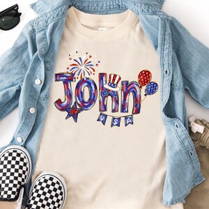 Puede incluir: Una camiseta color crema con el nombre "John" en rojo, blanco y azul, decorada con fuegos artificiales, globos y un sombrero patri&oacute;tico. La palabra "USA" est&aacute; debajo del nombre. La camiseta se combina con una chaqueta vaquera y zapatos a cuadros.