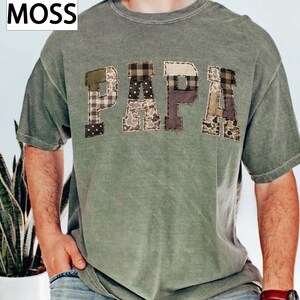 May include: A moss green t-shirt with the word "PAPA" appliqued across the chest. The letters are made of various patterned fabrics, including plaid, polka dots, and camouflage. The word "MOSS" is in the upper left corner.