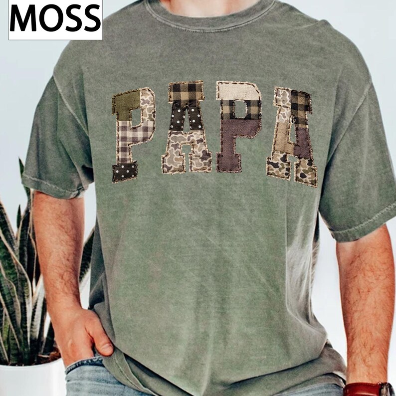 May include: A moss green t-shirt with the word "PAPA" appliqued across the chest. The letters are made of various patterned fabrics, including plaid, polka dots, and camouflage. The word "MOSS" is in the upper left corner.