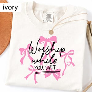 May include: Ivory-colored t-shirt with the text "Worship while you wait" in black script and pink bows. A gold necklace with a pendant is draped on the shirt. The shirt is made of a soft, comfortable material.