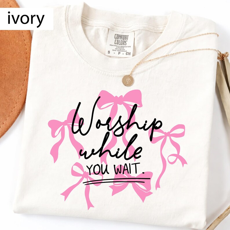 May include: Ivory-colored t-shirt with the text "Worship while you wait" in black script and pink bows. A gold necklace with a pendant is draped on the shirt. The shirt is made of a soft, comfortable material.
