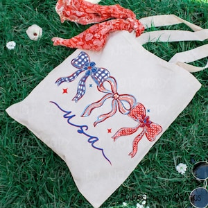 May include: A cream-colored tote bag with three decorative bows in red and blue, with the word "USA" in blue script. The bows have a gingham and polka dot pattern. A red patterned scarf and white straps are also visible.