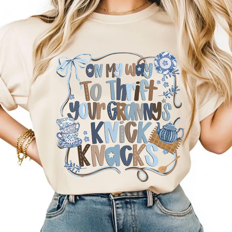 May include: Beige t-shirt with the phrase "On My Way To Thrift Your Granny's Knick Knacks" in blue and brown lettering. The design includes illustrations of teacups, a teapot, and floral accents, creating a vintage aesthetic.