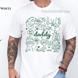 May include: White t-shirt with a green graphic design featuring the word "daddy" and various illustrations like a car, grill, and tools. The shirt also has text that says "Dad You Rock!" and "Best Dad Ever".