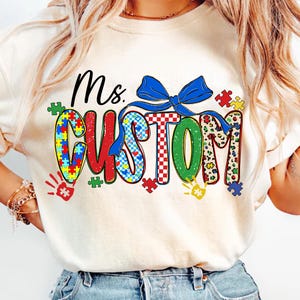 May include: A cream-colored t-shirt with the words "Ms. CUSTOM" in a colorful design. The word "CUSTOM" is decorated with puzzle pieces, a blue bow, and handprints. The shirt is a crew neck.