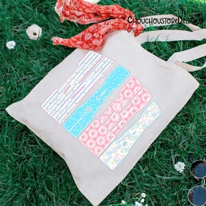 May include: A beige tote bag with the word "LOVE" in a colorful, patterned design. The letters are in blue, pink, and white, with floral and striped patterns. The bag has long, cream-colored handles and the text "CHOUCHOUSTOREDesigns" is visible.