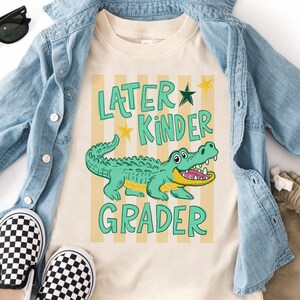May include: Cream-coloured t-shirt featuring a cartoon alligator and the text "Later Kinder Grader" in green. The alligator is light green with a yellow mouth. The shirt is paired with a denim jacket, checkered shoes, and khaki shorts.