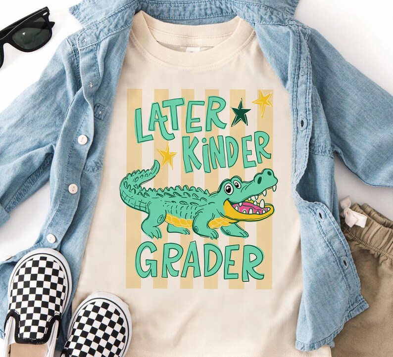 May include: Cream-coloured t-shirt featuring a cartoon alligator and the text "Later Kinder Grader" in green. The alligator is light green with a yellow mouth. The shirt is paired with a denim jacket, checkered shoes, and khaki shorts.