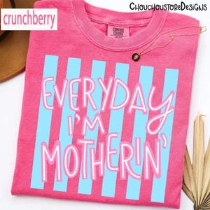 May include: A pink t-shirt with light blue vertical stripes and the text "EVERYDAY I'M MOTHERIN'" in white script. The shirt has a crew neck and short sleeves. The brand name "crunchberry" is in the top left corner.