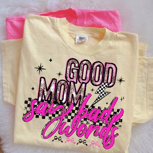May include: A light yellow t-shirt with the text "GOOD MOM say bad words" in pink and leopard print. A pink t-shirt is visible behind the yellow shirt. The design includes stars, a lightning bolt, and a checkered pattern.
