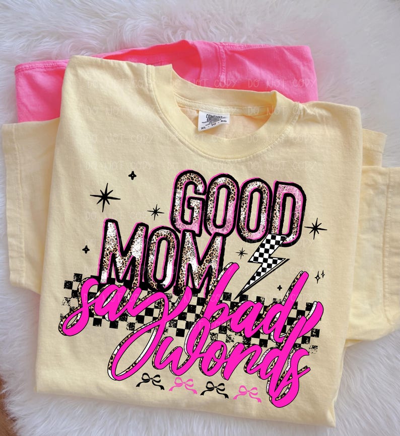May include: A light yellow t-shirt with the text "GOOD MOM say bad words" in pink and leopard print. A pink t-shirt is visible behind the yellow shirt. The design includes stars, a lightning bolt, and a checkered pattern.