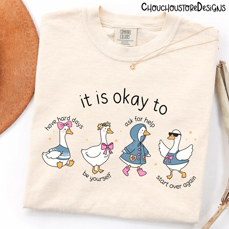 May include: Cream-colored t-shirt with the text "it is okay to" above four cartoon geese. Each goose is styled differently, with phrases like "have hard days" and "start over again" below them. The brand name "CHOUCHOUSTORE DESIGNS" is at the top.