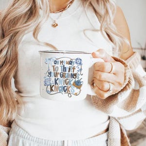 May include: White enamel camping mug with a black rim. The mug features the text "ON MY WAY TO THRIFT YOUR GRANNY'S KNICK KNACKS" in blue and brown lettering with decorative illustrations. A cozy accessory for coffee or tea.