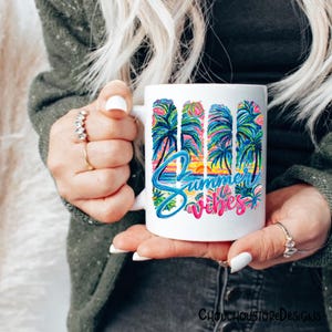 May include: White ceramic mug with a colorful "Summer Vibes" design. The design features palm trees, a sunset, and the words "Summer Vibes" in blue and pink. The mug is held by a person.