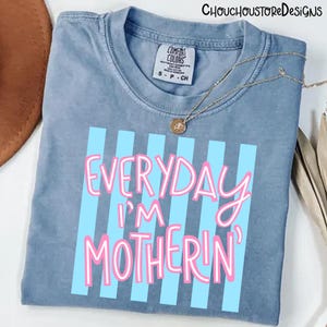 May include: A blue Comfort Colors t-shirt with the text "EVERYDAY I'M MOTHERIN'" in pink lettering with blue vertical stripes. A gold necklace with a pendant is draped on the shirt.