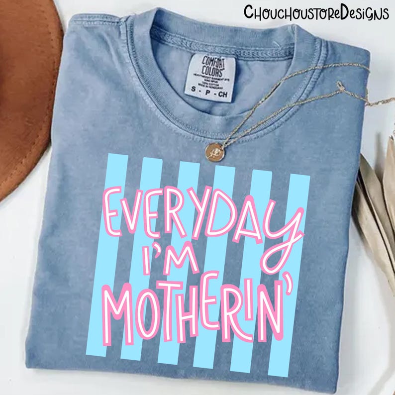 May include: A blue Comfort Colors t-shirt with the text "EVERYDAY I'M MOTHERIN'" in pink lettering with blue vertical stripes. A gold necklace with a pendant is draped on the shirt.