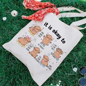 May include: A cream-colored tote bag with the text "it is okay to" and illustrations of cartoon Highland cows. The bag features the phrases "Have Hard Days", "Be Yourself", "Ask for Help", "Start Over Again", "Feel Your Feelings", and "Make Mistakes".