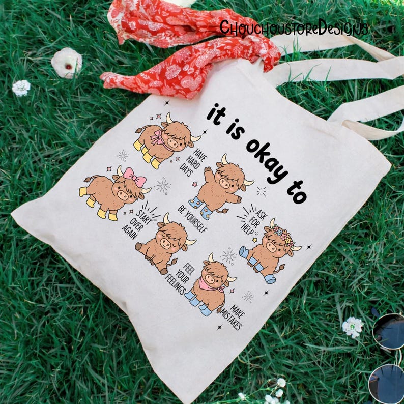 May include: A cream-colored tote bag with the text "it is okay to" and illustrations of cartoon Highland cows. The bag features the phrases "Have Hard Days", "Be Yourself", "Ask for Help", "Start Over Again", "Feel Your Feelings", and "Make Mistakes".