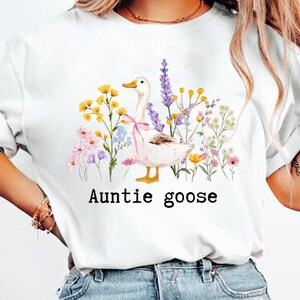 May include: White t-shirt featuring a watercolor illustration of a goose with a pink bow, surrounded by colorful flowers. The text "Auntie goose" is printed below the illustration in a black, sans-serif font.