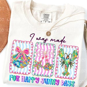 以下が含まれることがあります： 「I was made for happy sunny days」の文字が入ったクリーム色のTシャツ。貝殻、ヒトデと貝殻、ロブスターの3つのカラフルなフレームデザインが特徴です。金のネックレスがTシャツの上にかけられています。