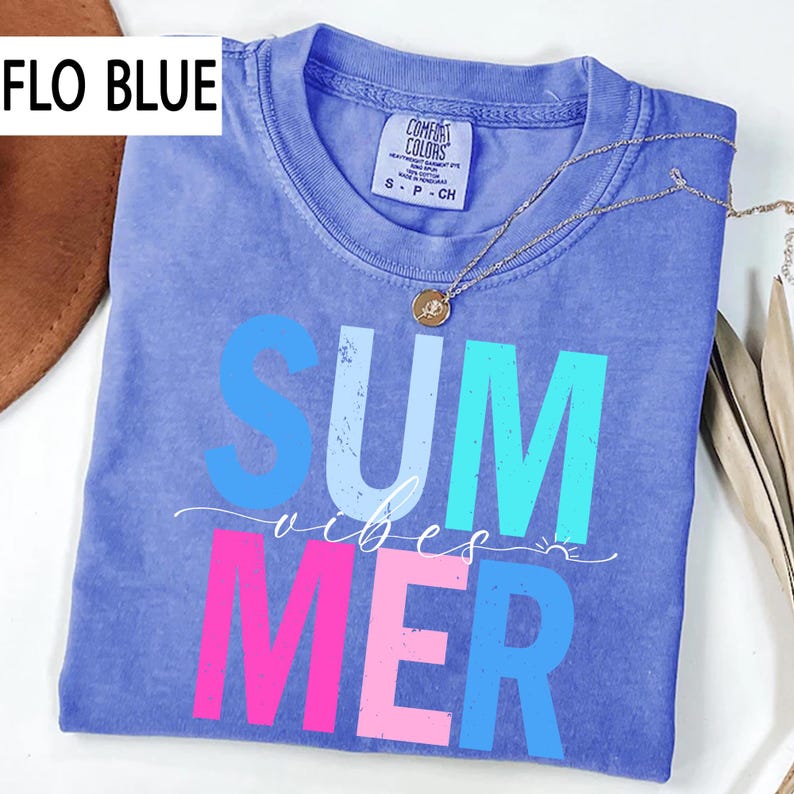 May include: A Flo Blue t-shirt with the words "SUMMER vibes" in blue, white, and pink. The shirt has a crew neck and short sleeves. A silver necklace with a small pendant is draped across the shirt.
