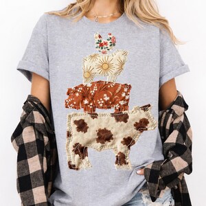 May include: A light gray t-shirt featuring a stacked animal design. The design includes a chicken, a sheep with sunburst details, a pig with floral patterns, and a cow with brown spots. The animals are made of fabric and stitched onto the shirt.