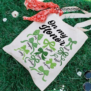 May include: A white canvas tote bag with the words "On my Honor" in black script. Green bows with words like "Caring", "Courageous", and "Fair" are printed on the bag. The bag has cream-colored handles.