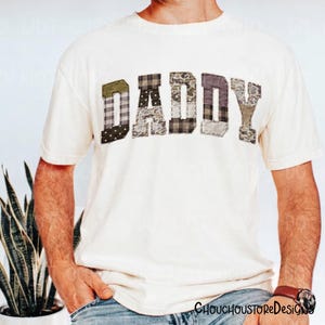 May include: Cream-colored t-shirt with the word "DADDY" across the chest. The letters are patchwork style, featuring various camouflage and plaid patterns in shades of green, brown, and gray. A casual, everyday tee.