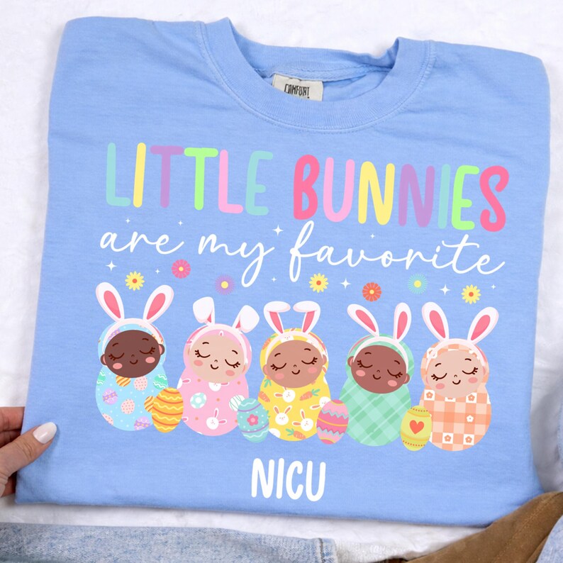 May include: A light blue sweatshirt with the text "LITTLE BUNNIES are my favorite" in colorful letters. The shirt features illustrations of five baby bunnies with Easter eggs and the word "NICU" at the bottom.
