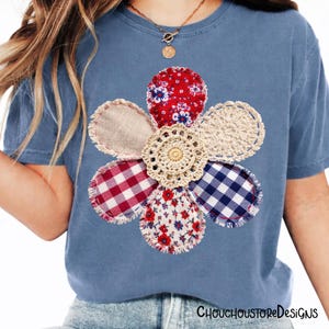 May include: A blue t-shirt with a large flower appliqu&eacute;. The flower is made from a variety of fabrics, including red floral, gingham, and crochet. The centre of the flower is a cream crochet doily. The shirt has a round neck and short sleeves.