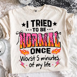 May include: Cream-colored t-shirt with the text "I TRIED TO BE NORMAL ONCE Worst 5 minutes of my life" in black, pink, and orange. The word "NORMAL" is in a pink checkered pattern with orange flames. The shirt has a crew neck and short sleeves.