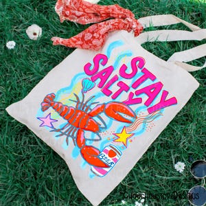 Puede incluir: Un bolso tote beige con las palabras "Stay Salty" en rosa y azul. El bolso presenta una langosta roja, una lata de cerveza y gr&aacute;ficos de estrellas. El bolso tiene asas color crema y est&aacute; sobre un lecho de hierba verde.