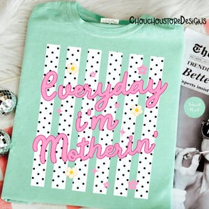 May include: A pale green t-shirt featuring the pink text "Everyday I'm Motherin". The design incorporates white vertical stripes with black polka dots, and small flower and star motifs.