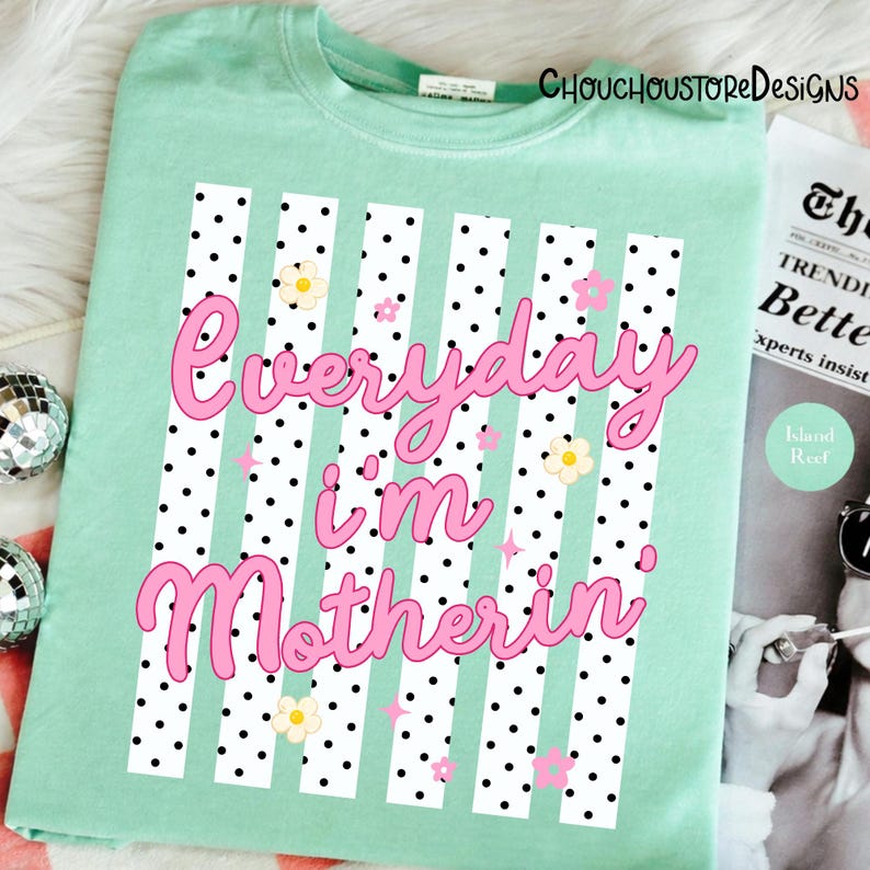 May include: A pale green t-shirt featuring the pink text "Everyday I'm Motherin". The design incorporates white vertical stripes with black polka dots, and small flower and star motifs.