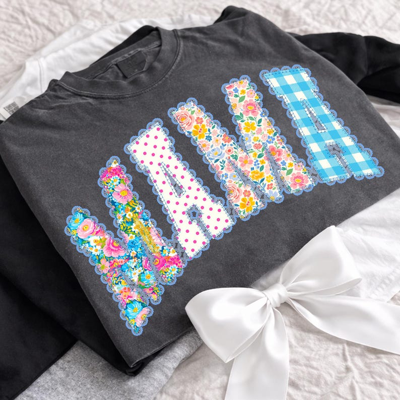 May include: Charcoal grey sweatshirt featuring the word "NANA" in decorative letters. The letters are adorned with floral, polka dot, and gingham patterns, edged with a scalloped trim. A white satin bow is visible.
