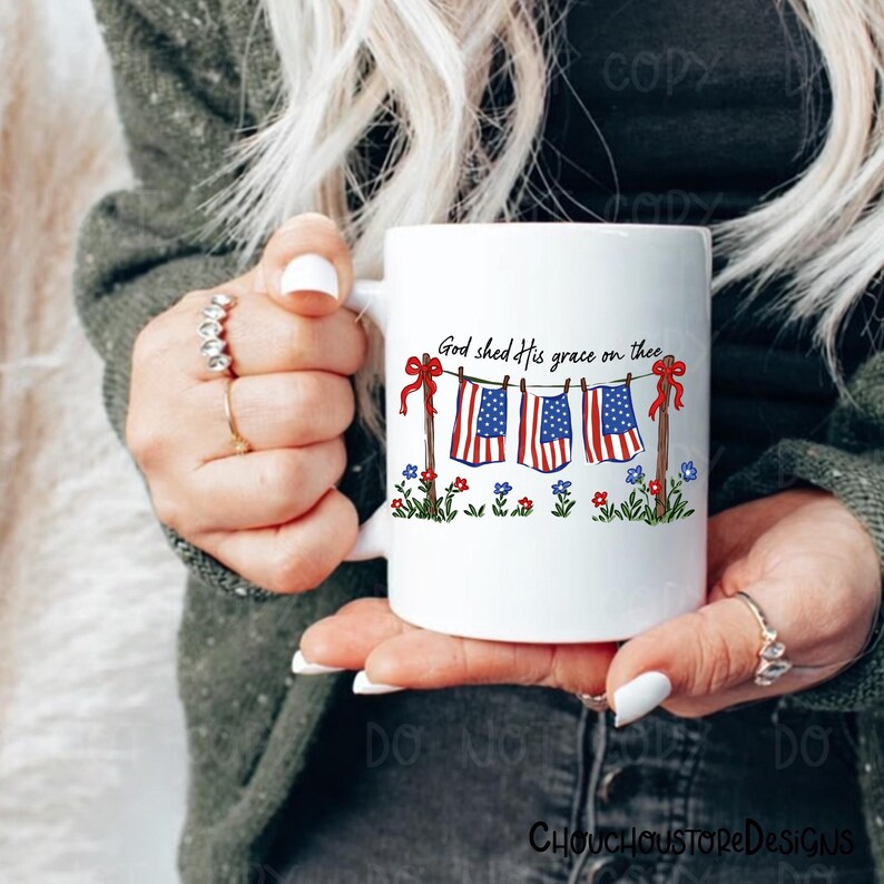 May include: White ceramic mug featuring a patriotic design. The mug displays three American flags hanging on a clothesline, with the text "God shed His grace on thee." Red bows and floral accents complete the design.