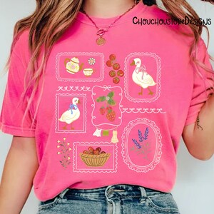 May include: A pink t-shirt with various framed illustrations. The illustrations include a teapot, teacup, goose with a bow, strawberries, and a basket of fruit. The text "ChouchouStoreDesigns" is printed at the top right.