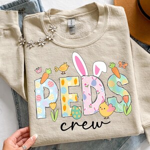 May include: Beige crewneck sweatshirt with the text "REDS crew" in colorful, Easter-themed lettering. The design includes bunny ears, carrots, eggs, birds, and flowers, perfect for the spring season.