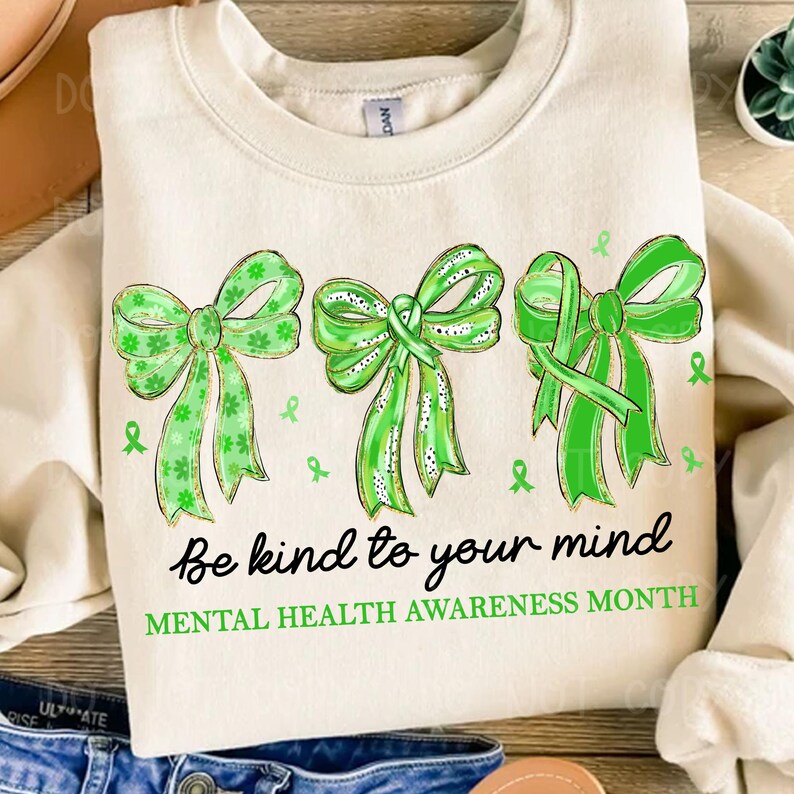 May include: Cream-colored sweatshirt featuring three green bows with varying designs. The text "Be kind to your mind" and "MENTAL HEALTH AWARENESS MONTH" are printed below the bows. Small green ribbons are scattered around the bows.