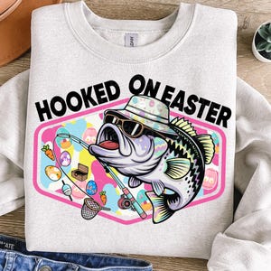 May include: Light gray sweatshirt with the text "HOOKED ON EASTER" above a cartoon bass wearing sunglasses and a bucket hat. The bass is holding a fishing rod with Easter eggs and carrots. The design is set in a pink and blue frame.