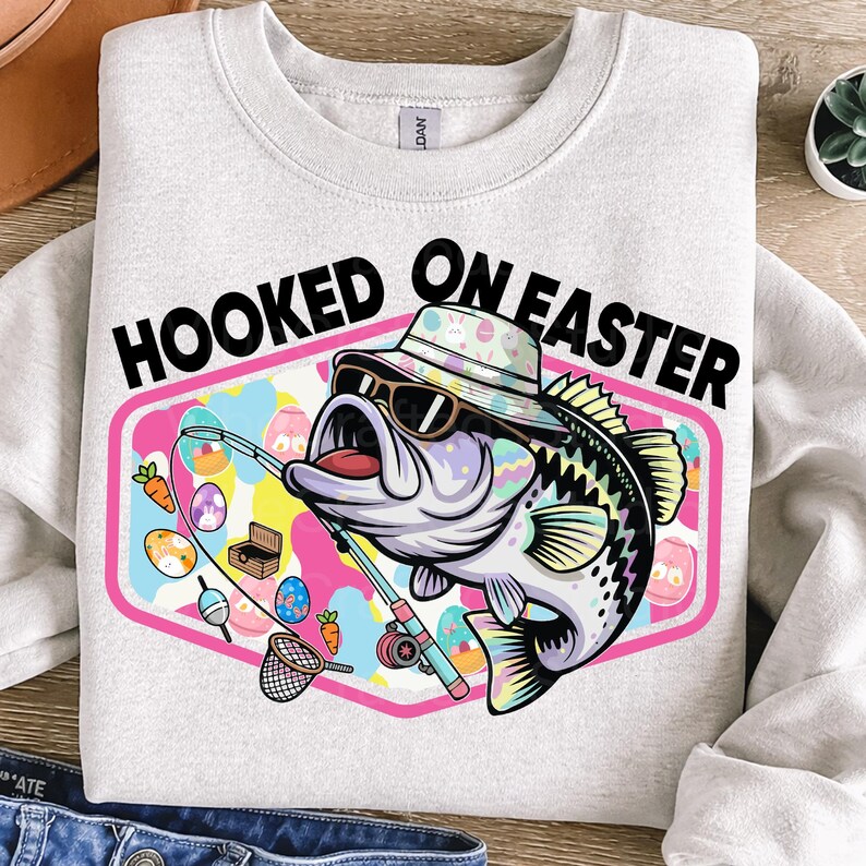 Pode incluir: Camisola cinza claro com o texto "HOOKED ON EASTER" acima de um desenho de baixo de desenho animado usando &oacute;culos de sol e um chap&eacute;u de balde. O baixo est&aacute; segurando uma vara de pescar com ovos de P&aacute;scoa e cenouras. O design est&aacute; em uma moldura rosa e azul.
